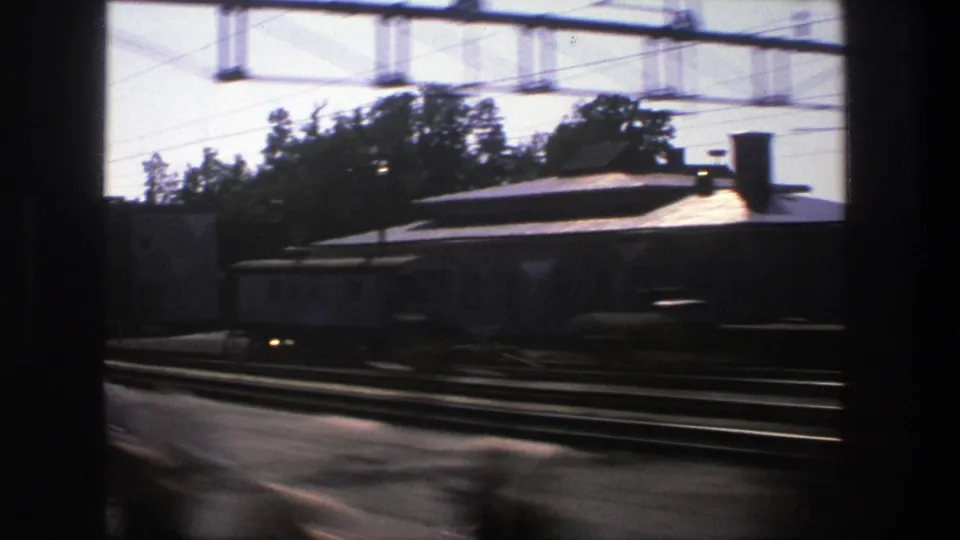 1975 Stockholm rail scene from a moving train, showing tracks, a low station building, trees, and overhead power equipment.