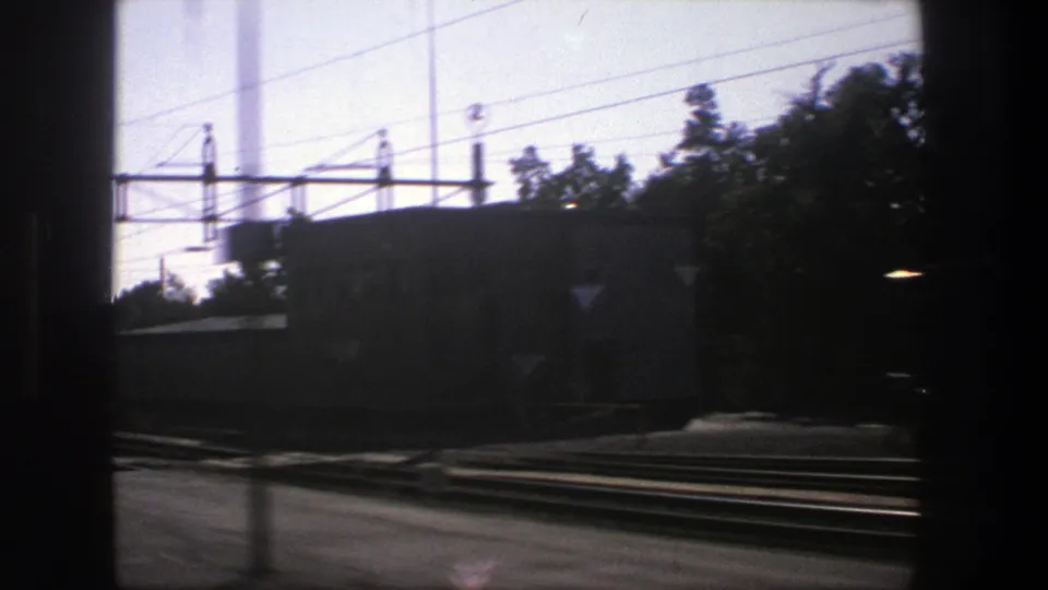 End frame of the 1975 Stockholm rail pass-by, with dark window framing, tracks, overhead lines, trees, and a station-side building.