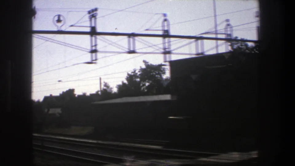 1975 Stockholm rail corridor viewed from a moving train, with tracks, overhead wires, utility masts, trees, and dark window edges.
