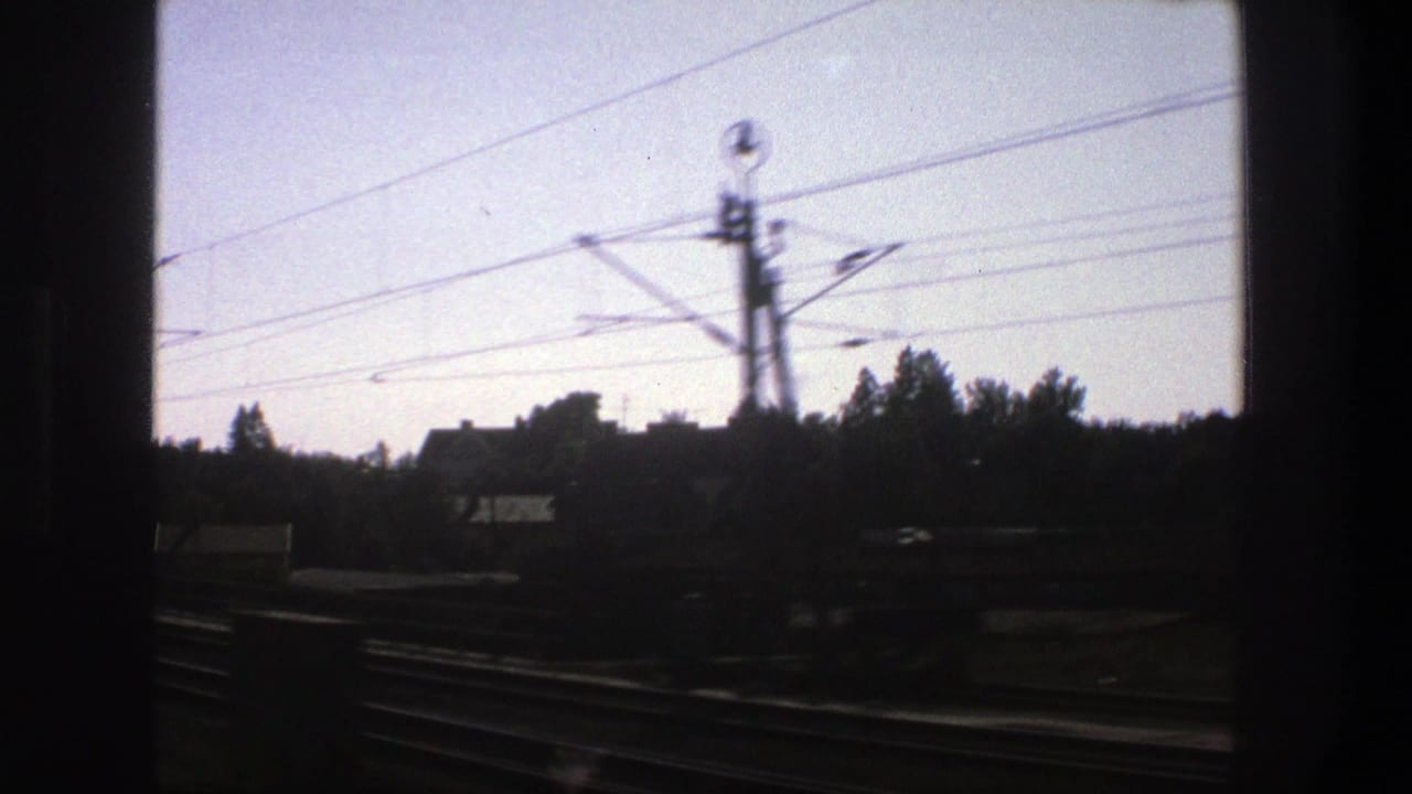 Moving 8mm view in Stockholm, Sweden, showing overhead rail wires, trackside poles, trees, rooftops, and a dark window divider.
