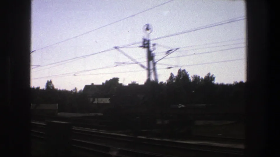Moving 8mm view in Stockholm, Sweden, showing overhead rail wires, trackside poles, trees, rooftops, and a dark window divider.