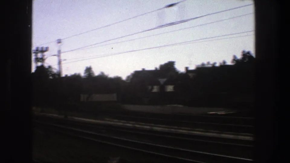 Stockholm train-window footage from 1975 with blurred tracks, overhead wires, dark window edges, trees, and low structures beyond the railway.