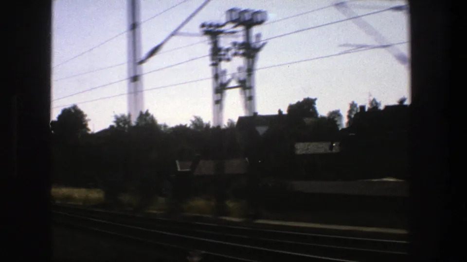 Final 1975 Stockholm rail view with multiple tracks, overhead electric wires, a trackside utility structure, trees, rooftops, and dark film edges.