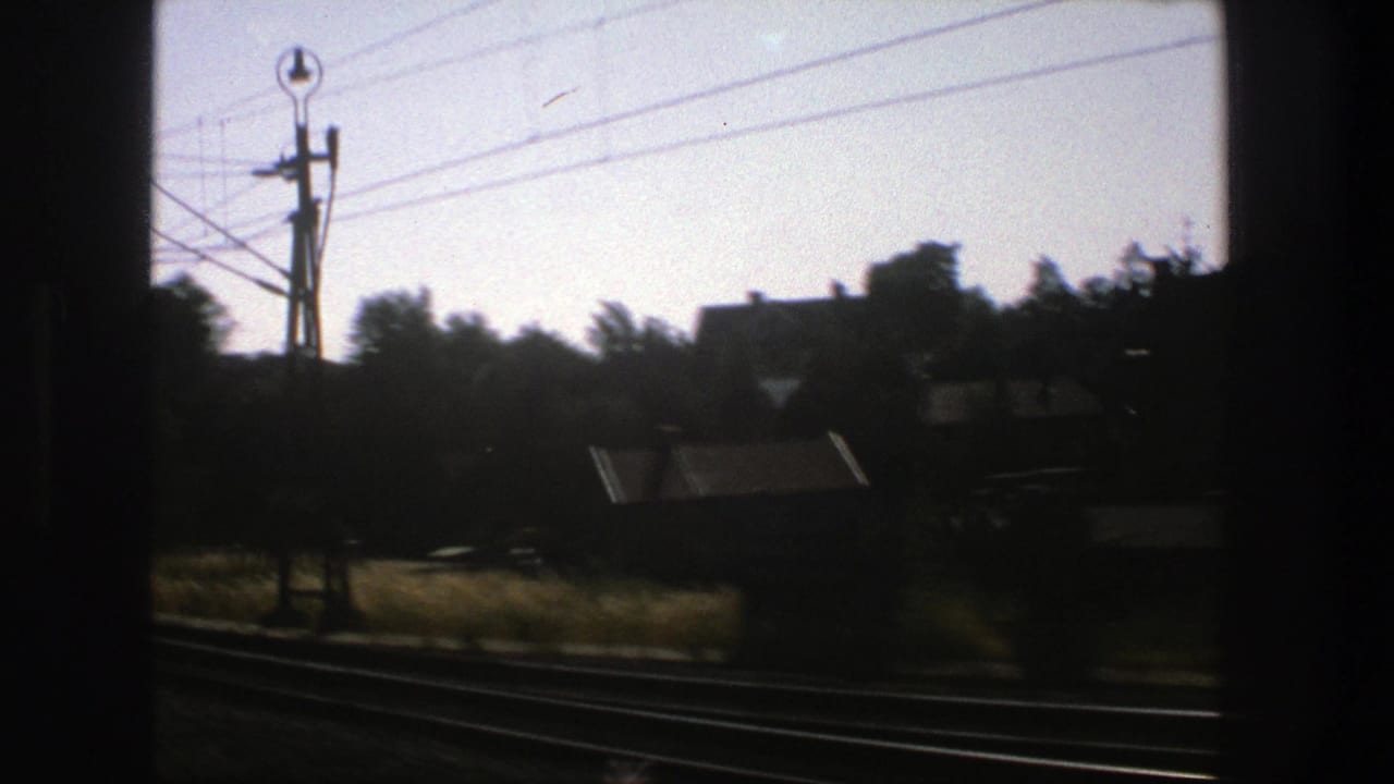 Dark train-window view of rail tracks, houses, trees, and overhead power lines.