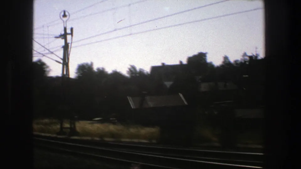 1975 Stockholm 8mm frame from a moving train, with dark window bars, rail tracks, houses, trees, and utility lines.