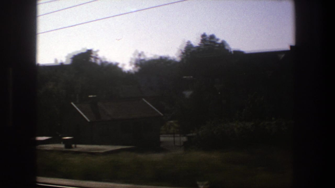 Moving 8mm train view in Stockholm, showing a dim rail-side neighborhood with small buildings, trees, tracks, and overhead lines.