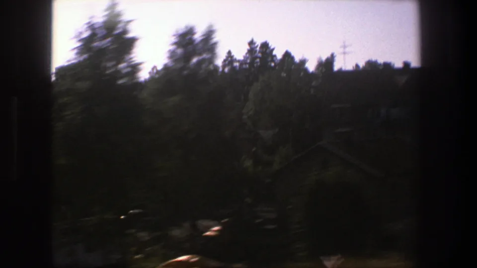 Dark, motion-blurred 1975 Stockholm train-window frame with trees, rooftops, a utility mast, and shadowed foreground.