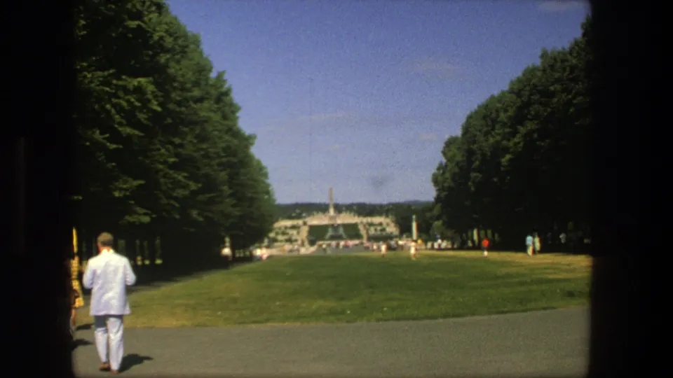 1975 Stockholm park scene with a man in a light suit on a path, lawns, tree rows, visitors, blue sky, and a distant landmark-like structure.