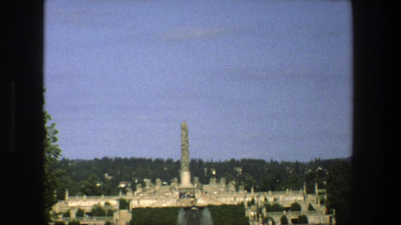 Window-framed view of a tall monument, park grounds, trees, sky, and distant visitors.