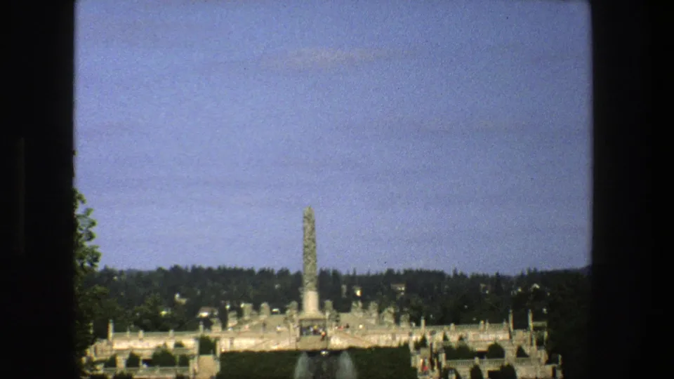 1975 Stockholm home-movie still with a tall monument, fountain spray, formal grounds, trees, and tiny visitors in the distance.