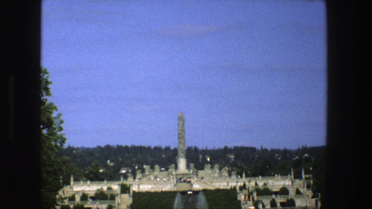 1970s 8mm travel frame showing a wide monument park view with a tall central monolith, terraces, greenery, and dark window-like borders.