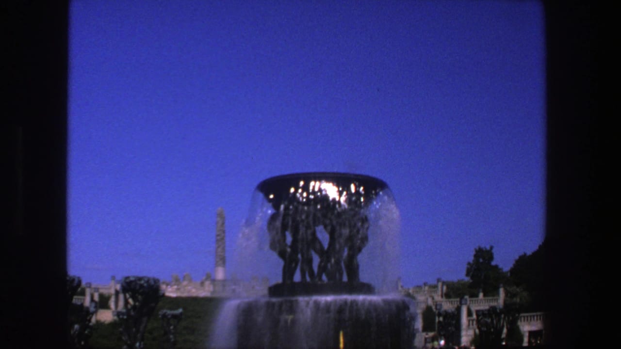 Large dark sculptural fountain with water flowing, a tall monument behind it, and blue sky.