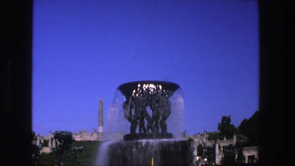 1970s 8mm travel frame showing a fountain with water cascading before terraces, a tall monument, greenery, and small park visitors.