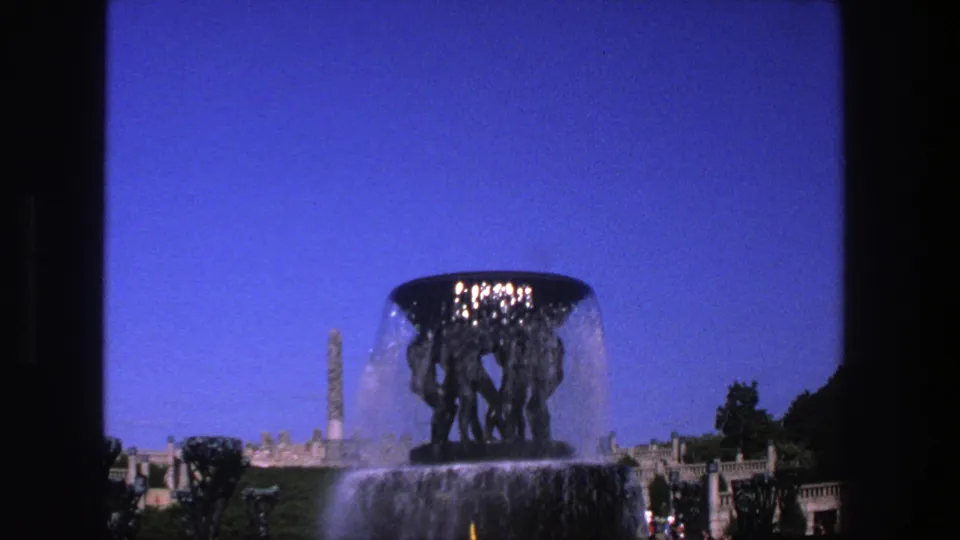 1970s 8mm frame of a monumental fountain with flowing water, formal park terraces, a tall background monument, and dark film-frame edges.