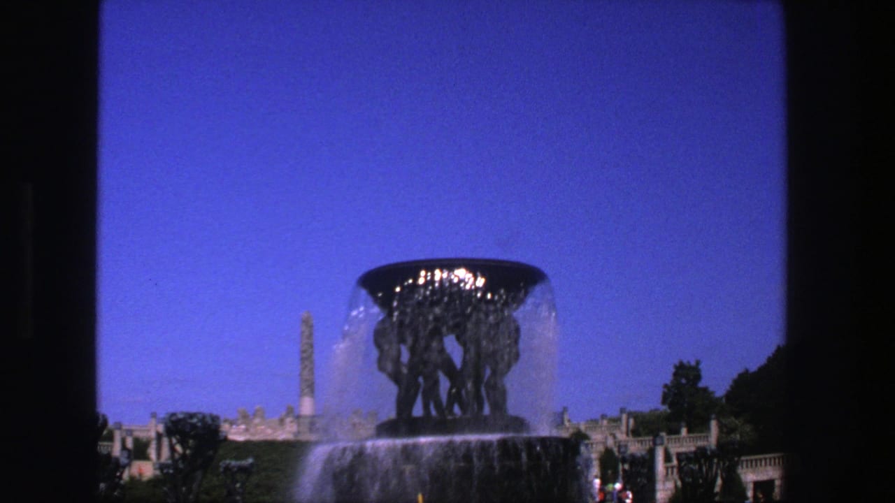Sculptural fountain spraying water beneath a blue sky with an obelisk-like monument behind it.