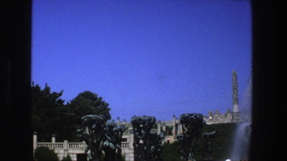 1975 8mm frame identified as Stockholm, shifting to a wider park view with fountain structures, trees, railings, blue sky, and water at the edge.