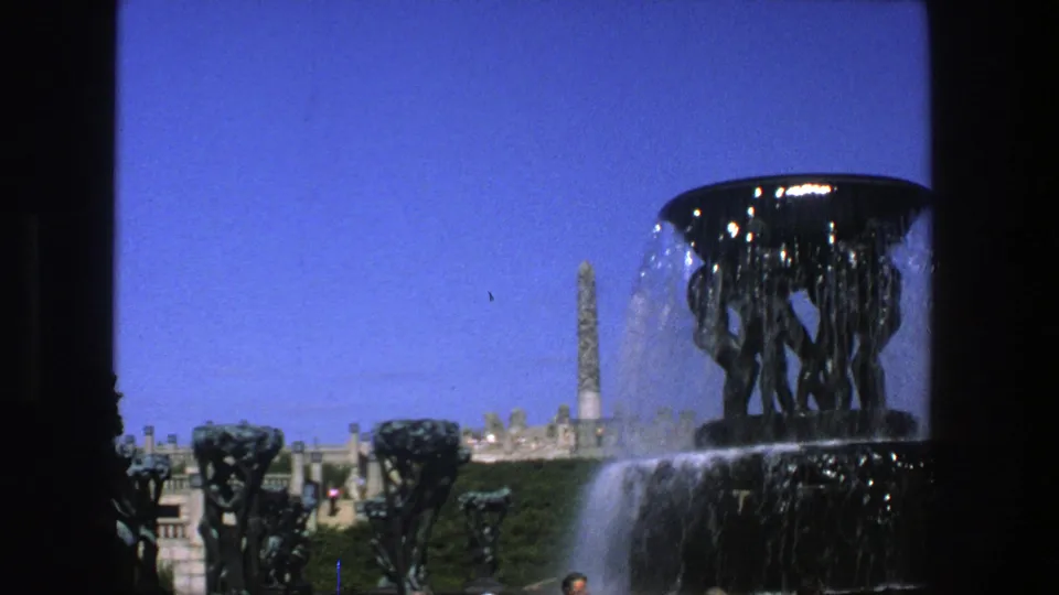 1975 Stockholm frame with dark fountain sculptures, water spray, a distant obelisk-like monument, terrace structures, and a small visitor at bottom edge.