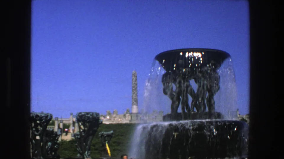 1975 Stockholm 8mm still showing a dark fountain basin pouring water, bronze figures, park greenery, stone terrace, and distant obelisk-like monument.