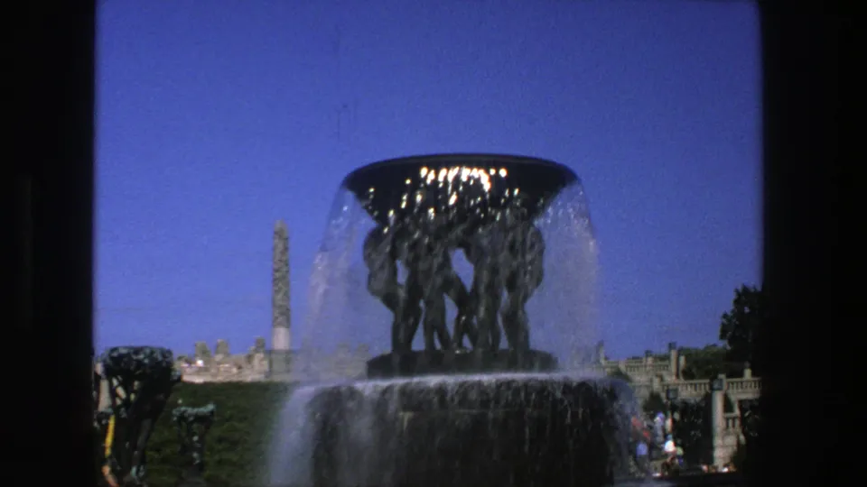 1975 Stockholm 8mm frame with the fountain centered, water flowing over the sculpture, and the obelisk visible beyond the park.