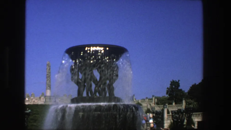 1975 Stockholm 8mm frame showing the fountain from a visitor viewpoint, with water spray, the obelisk, buildings, and people in the background.