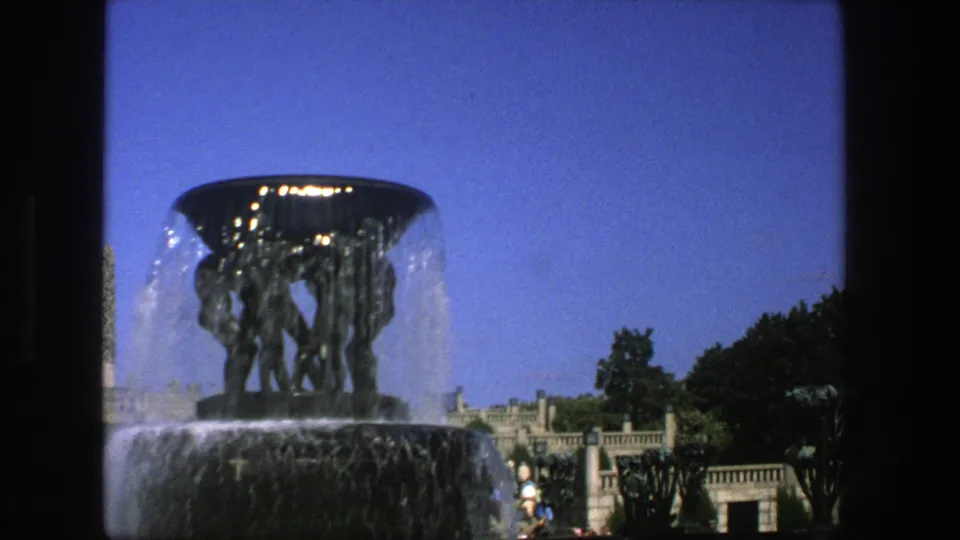 1975 Stockholm 8mm frame of the public fountain and sculpture, with trees, balustrade, obelisk, and distant visitors under blue sky.