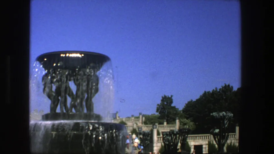 1975 Stockholm 8mm frame showing a large sculptural fountain, blue sky, trees, stonework, and distant park visitors.