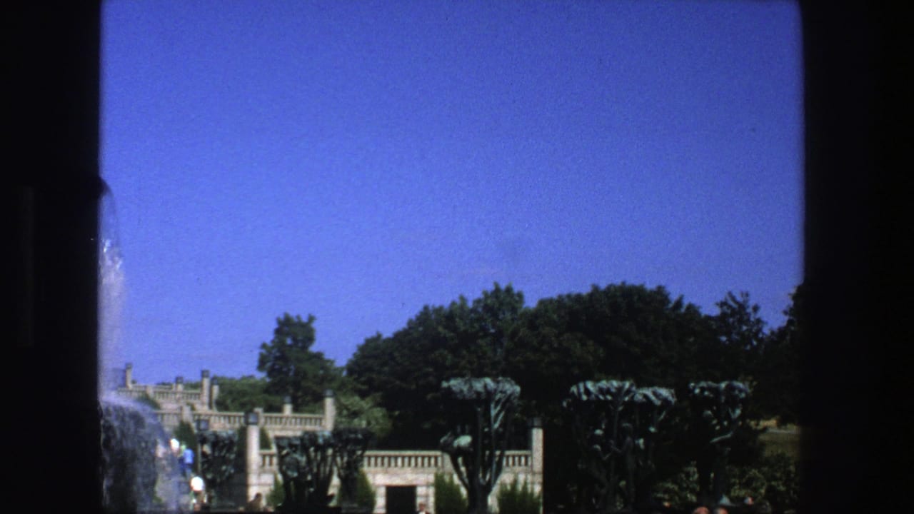 Wide park view with a stone fountain, trees, clipped plantings, and blue sky.