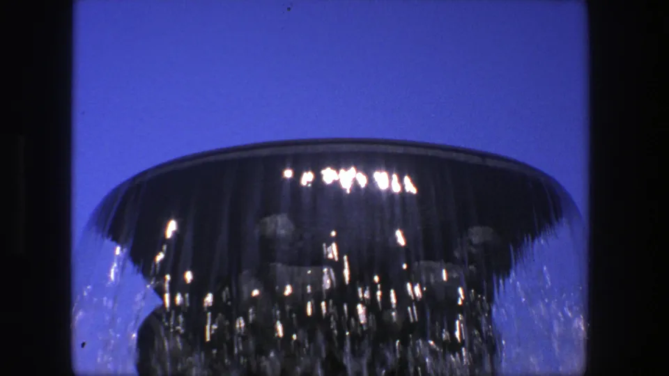 A close 8mm frame from Stockholm shows water sheeting over a dark stone fountain bowl, with saturated sky and bright reflected highlights.