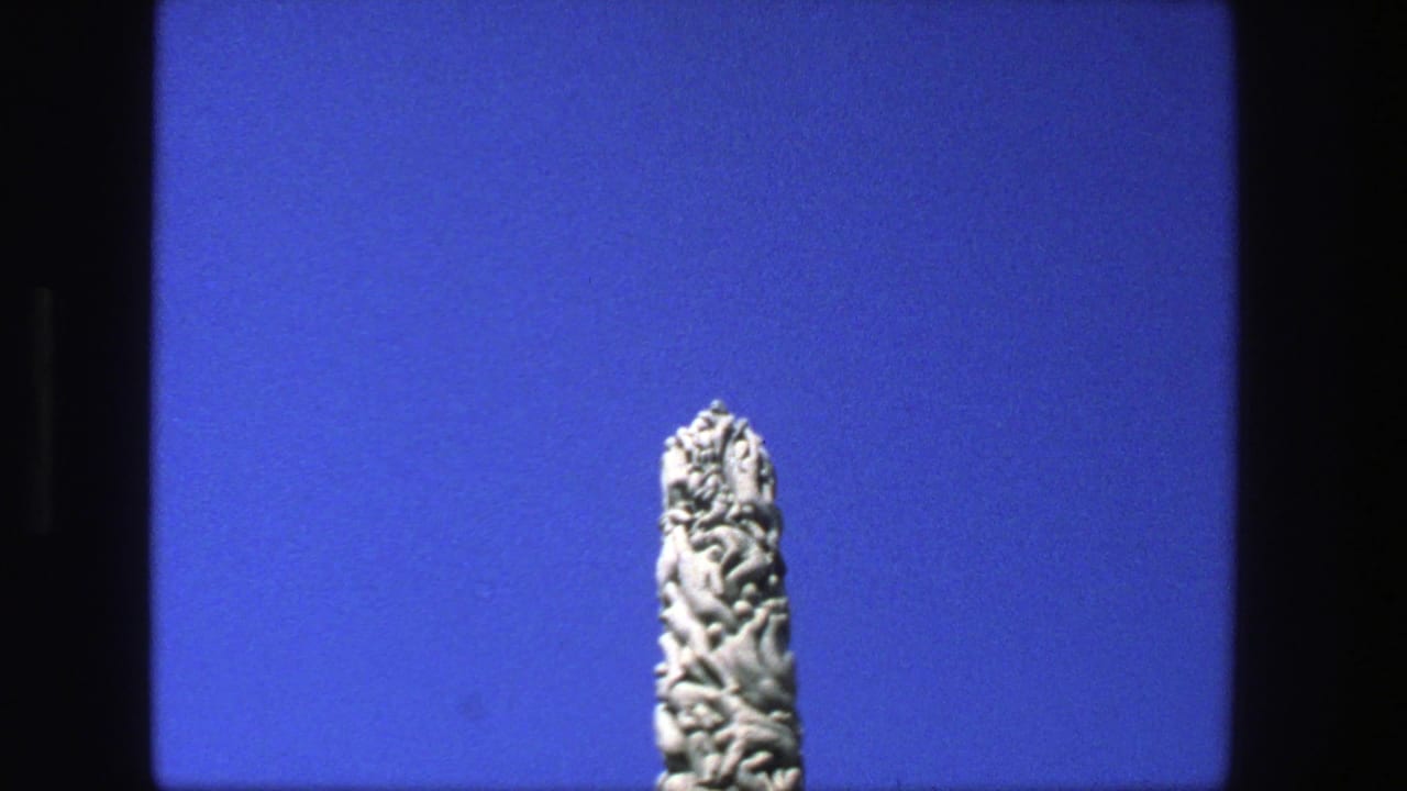 1975 Stockholm 8mm frame with the upper section of an intricately carved stone obelisk against blue sky.