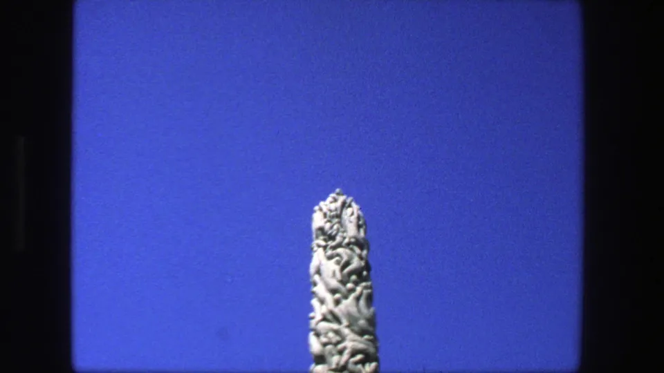 1975 Stockholm 8mm frame with the upper section of an intricately carved stone obelisk against blue sky.