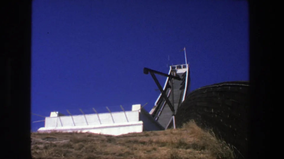1975 Stockholm 8mm view of a white hilltop installation with antenna equipment, dark railing, dry grass, and clear blue sky.