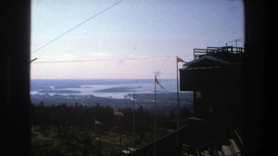 1975 Stockholm 8mm frame showing a distant archipelago view, flags in the foreground, and a dark observation deck on the right.