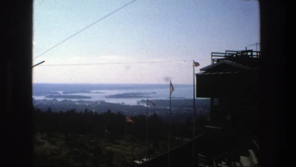 1975 Stockholm 8mm view across water and islands from a hilltop lookout, with flags and a dark wooden platform in silhouette.