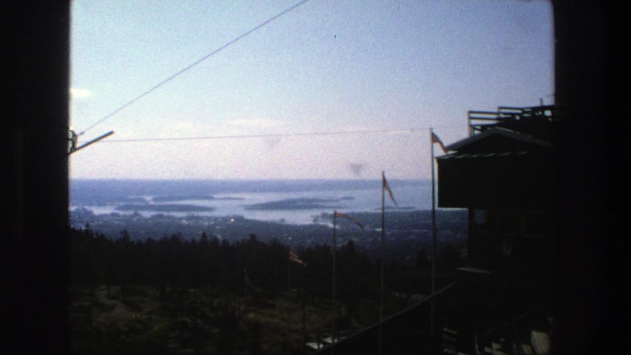 1975 Stockholm home-movie view from a high vantage point, with a cable line, dark lift structure, forest, and distant waterways.