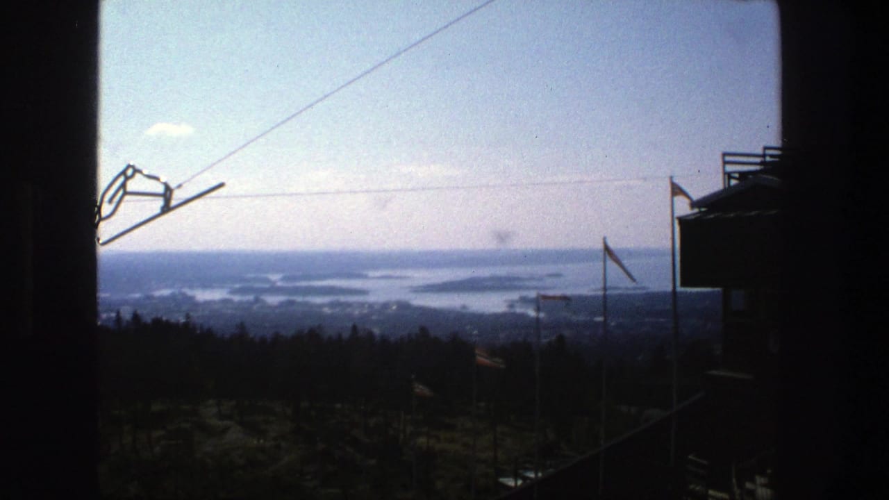 Cable hardware hangs against a pale sky over the Stockholm-area forest and distant archipelago, seen from an elevated home-movie viewpoint.