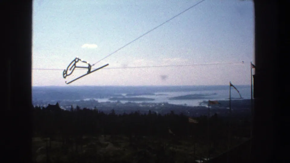 Stockholm-area high vantage view showing cable apparatus, conifer treetops, a small structure edge, flags, and distant blue waterways.