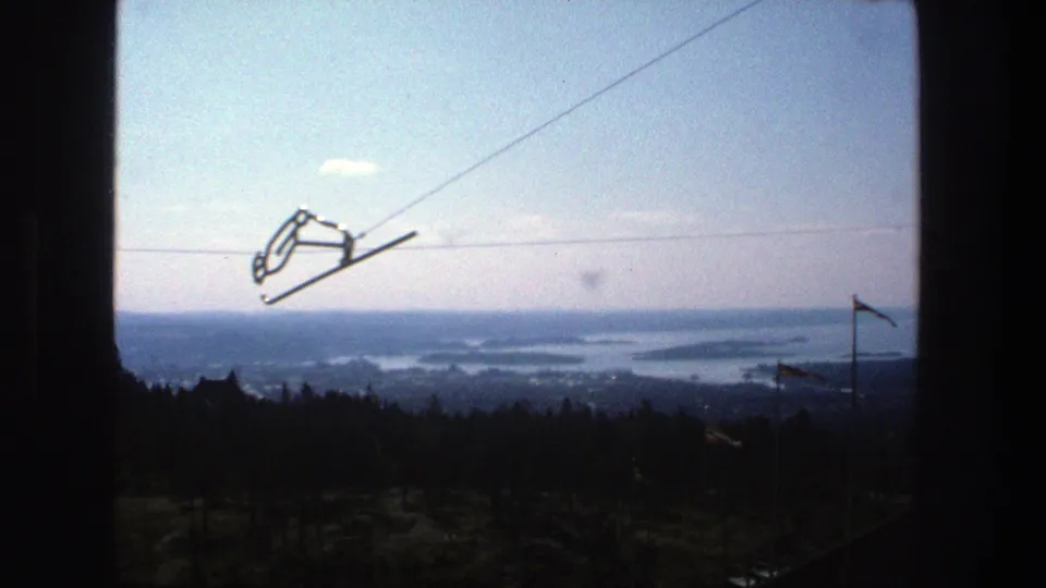 1975 Stockholm home-movie frame showing a cable car line silhouetted above forest and distant archipelago waters.