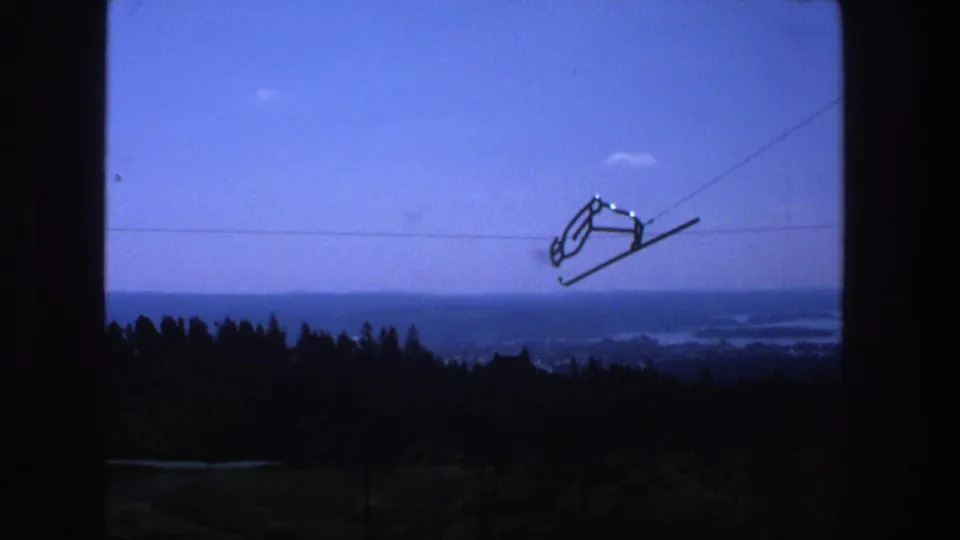 1975 Stockholm home-movie frame showing a ski-lift chair and cable silhouetted over trees, waterways, and hazy city distance.