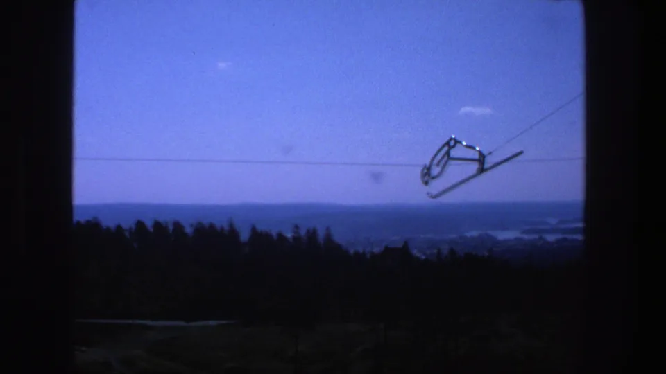 1975 8mm frame from Stockholm with a ski-lift chair on cables above a shadowed tree line and distant waterways.