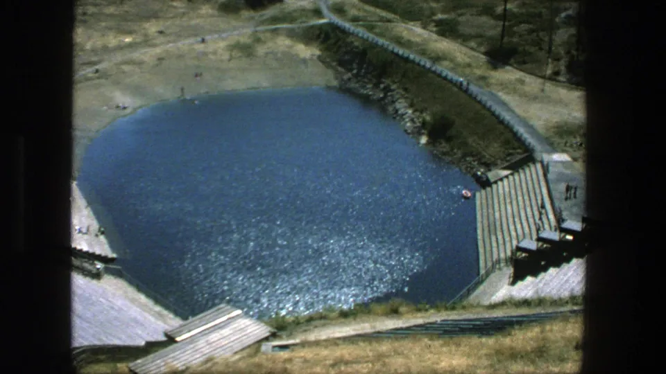 1975 Stockholm 8mm view looking down at a circular outdoor pool, concrete deck, steps, paths, grassy banks, and very small people.