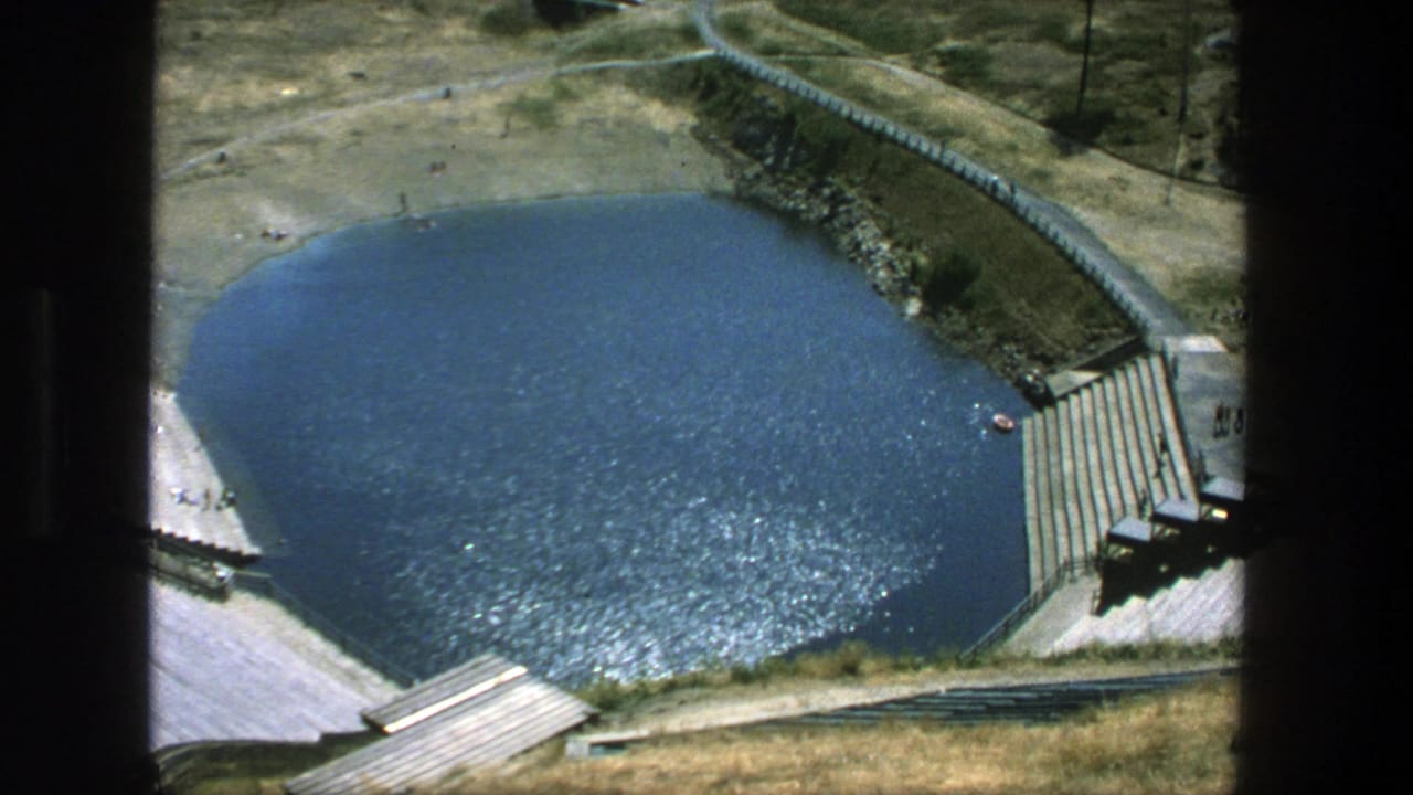 Elevated view of a blue outdoor pool with concrete steps, dry grass, and small people nearby.