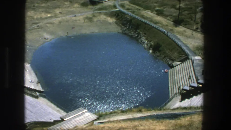 1975 Stockholm home-movie view of an outdoor pool from above, with blue water, steps, paths, and people visible at a distance.