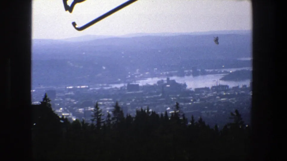Hazy 1975 Stockholm panorama from a wooded high point, showing water, city forms, distant hills, and part of a ski-jump structure above.