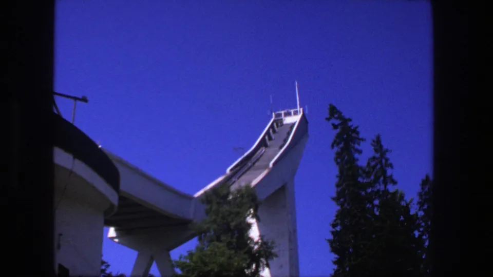 Low-angle 1975 Stockholm shot of a ski-jump structure rising beside tall trees under a saturated blue sky.