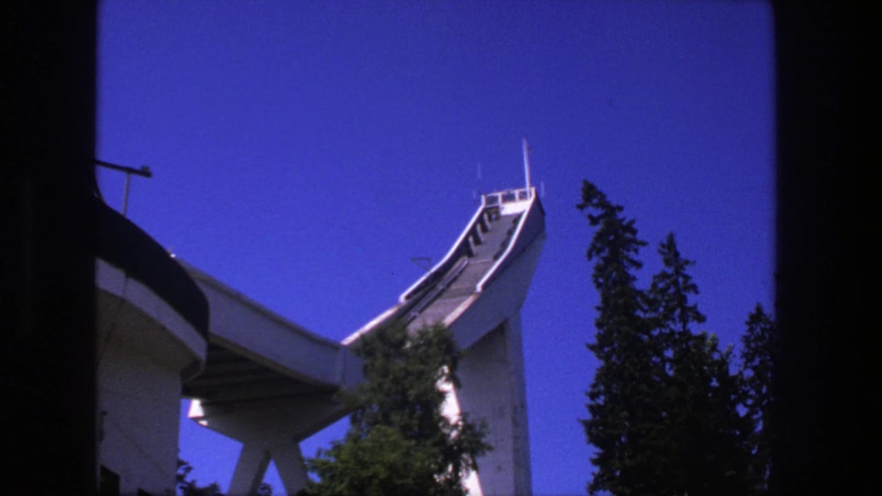 Concrete ski jump rising above trees against a blue sky.