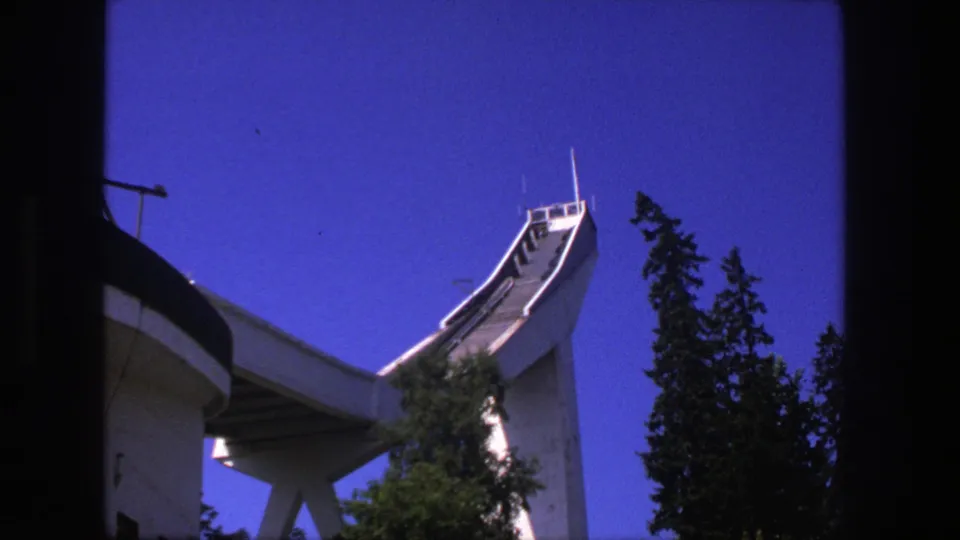 Stockholm 1975 8mm frame of an unfinished concrete ski jump viewed from below, with tree silhouettes and vivid blue sky.