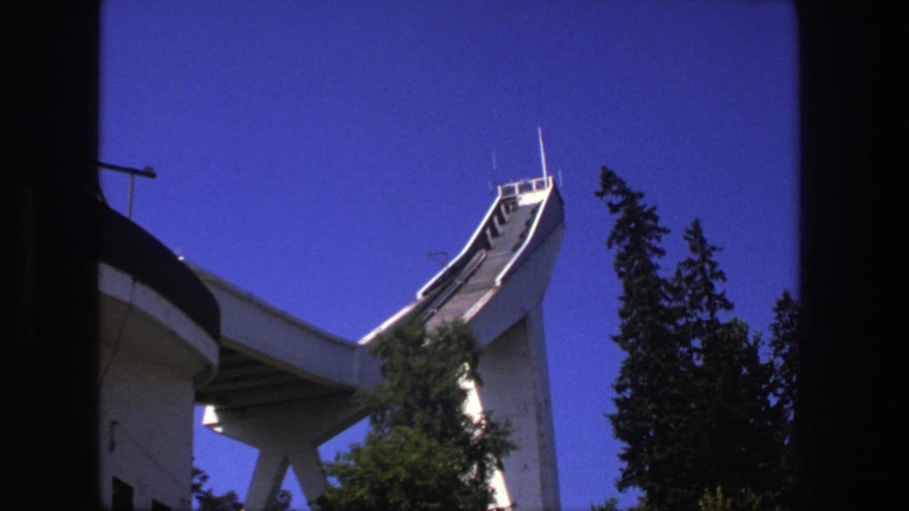 1975 8mm frame showing a white ski-jump structure rising above dark evergreen trees under a saturated blue sky.