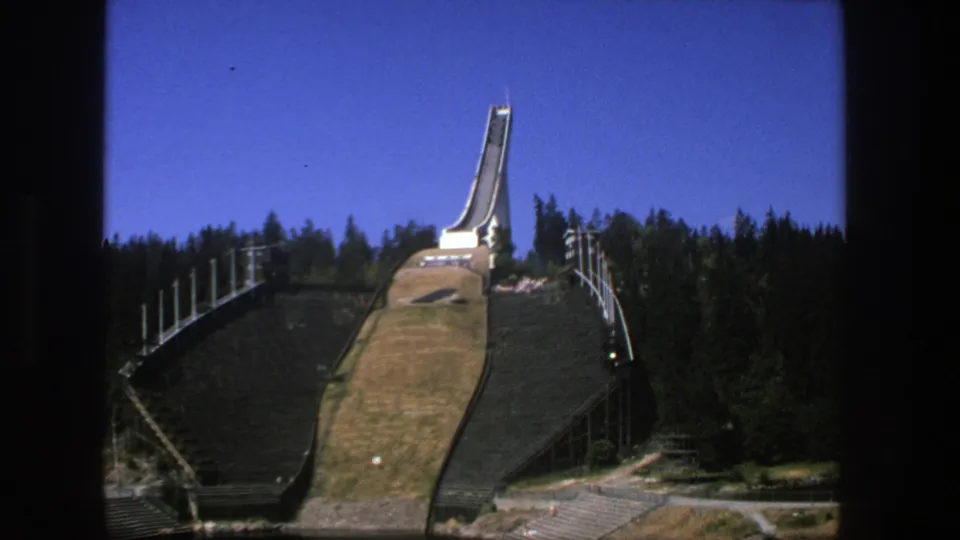 Wide 8mm view of the ski-jump landing slope and tower set among evergreen trees under clear blue sky.