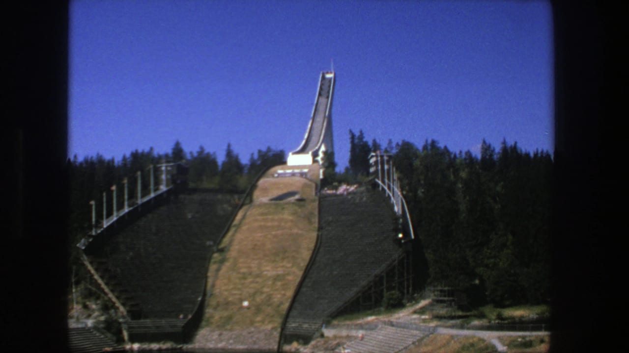 1975 8mm view of a large ski jump structure above forested hillside stands, seen from across a waterside foreground.