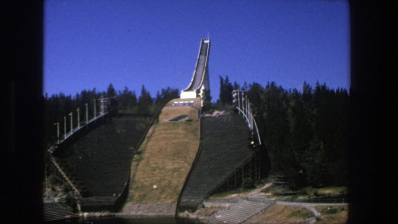 Vintage 8mm frame of the stated Stockholm ski jump setting, with forest, terraces, paths, and water visible below.
